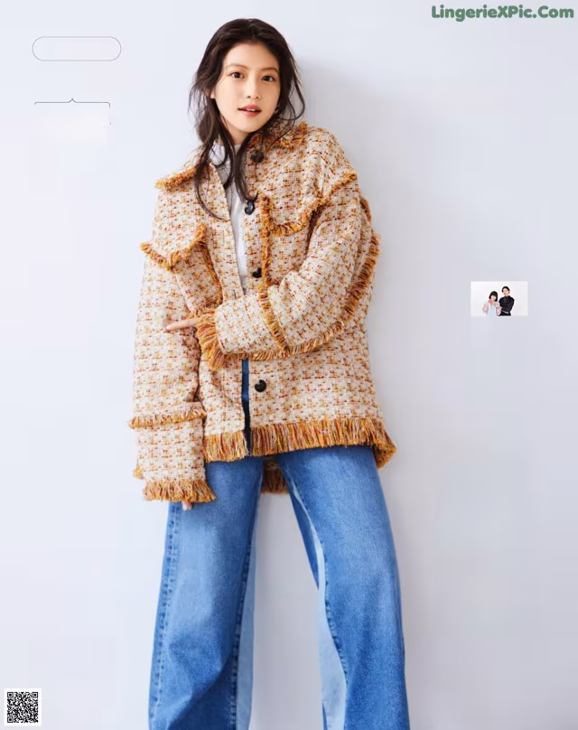 A woman standing in front of a white wall wearing a jacket and jeans.