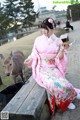 A woman in a pink kimono sitting on a bench next to a deer.